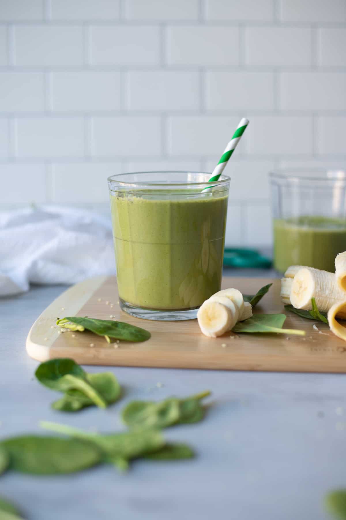 Green smoothie in a glass sitting on a wooden cutting board next to sliced bananas on a kitchen counter.