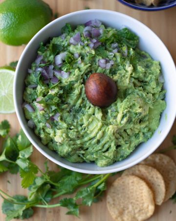 Top down view of guacamole in a bowl with chips, cilantro, and a lime around it.